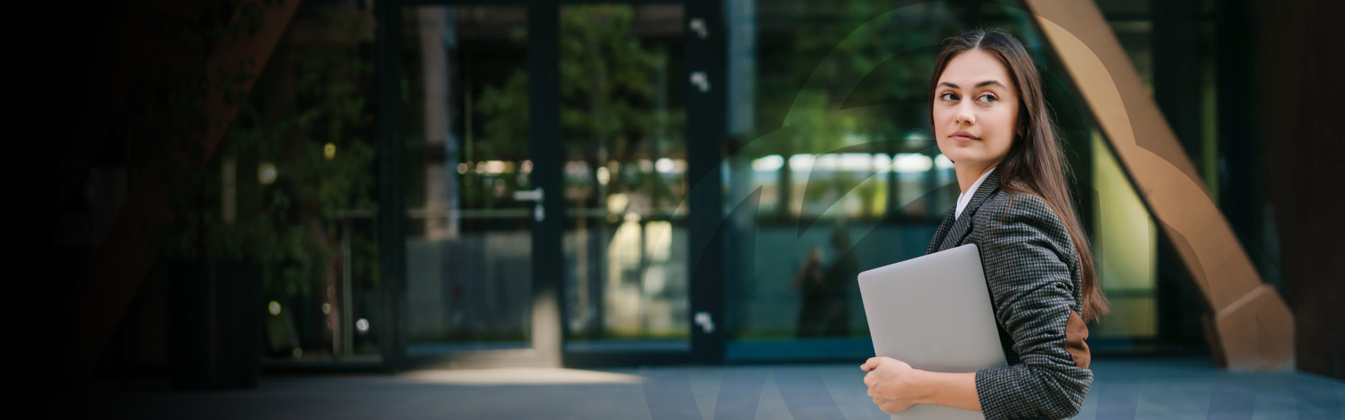 A businesswoman in a blazer glancing over her shoulder while holding a laptop, standing outside a glass office building.