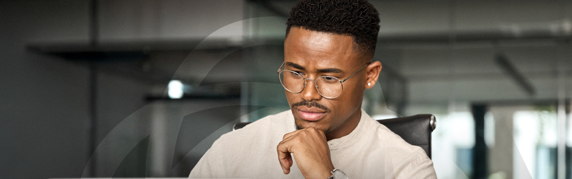 A man in round glasses sits at a desk in a modern office, resting his chin on his hand as he studies something in front of him.
